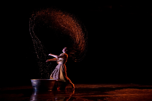 Lone female performer on darkened stage in dramatic lighting throwing her wet hair back in a high arc.